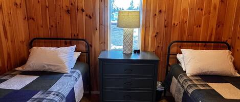Habitación tipo cabaña con dos camas, una mesita de noche con lámpara y una ventana con vista a árboles.