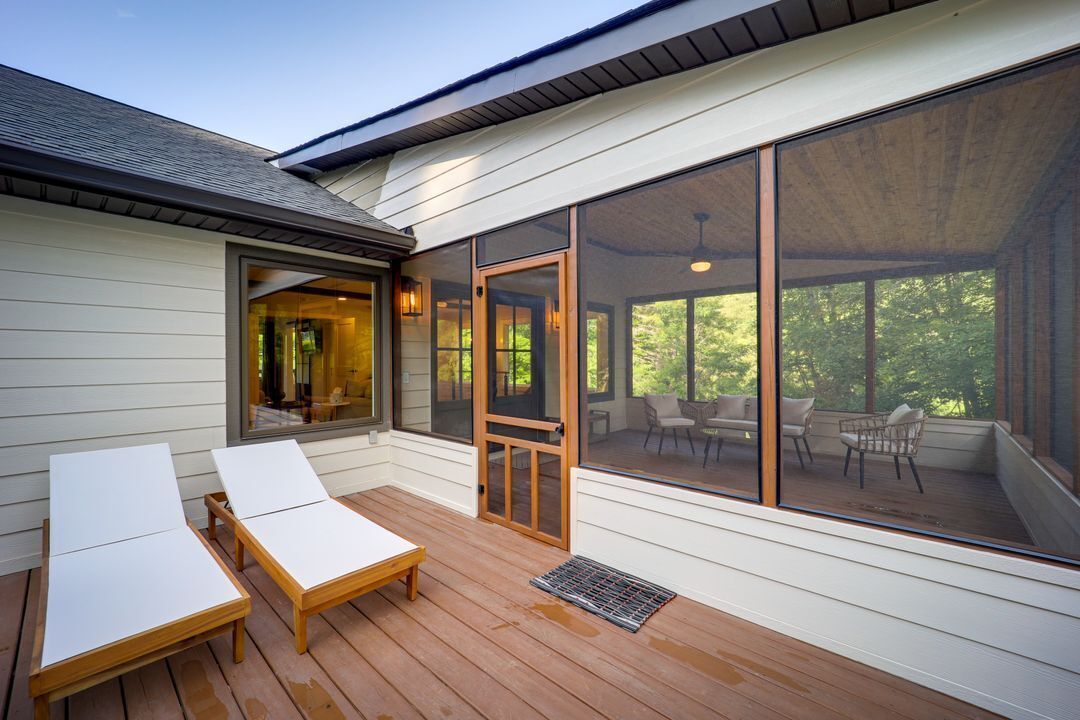 A covered outdoor area with two white lounge chairs, a wooden table, and a door leading to a patio with outdoor furniture.