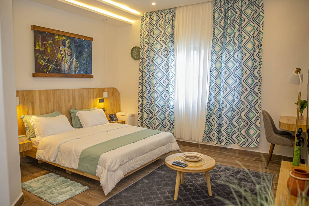 Una moderna camera d'hotel con un letto con testiera in legno, un comodino con lampada, un tavolino rotondo e tende a motivi.