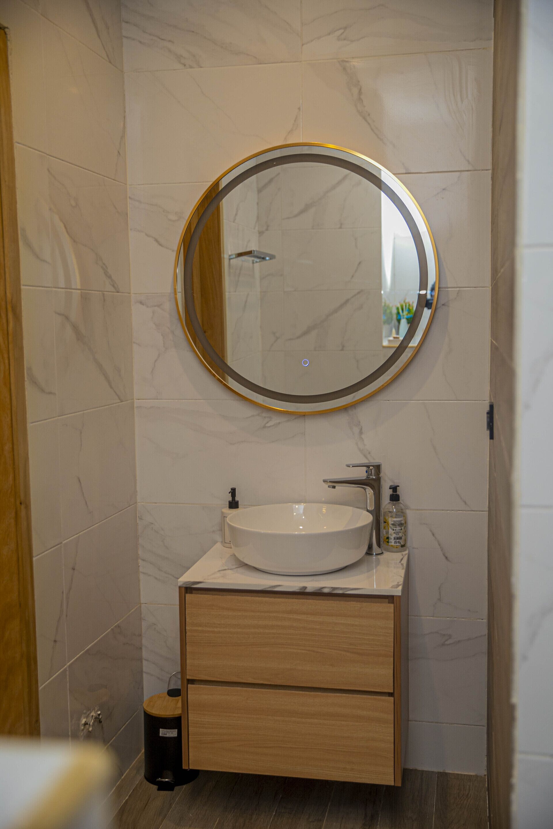 Un baño con pared de azulejos de mármol, un espejo redondo con marco dorado, un lavamanos blanco y un tocador de madera con dos cajones.