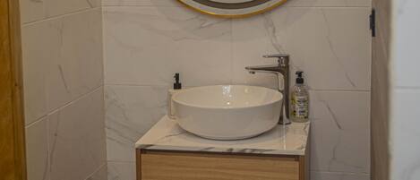 Un baño con pared de azulejos de mármol, un espejo redondo con marco dorado, un lavamanos blanco y un tocador de madera con dos cajones.