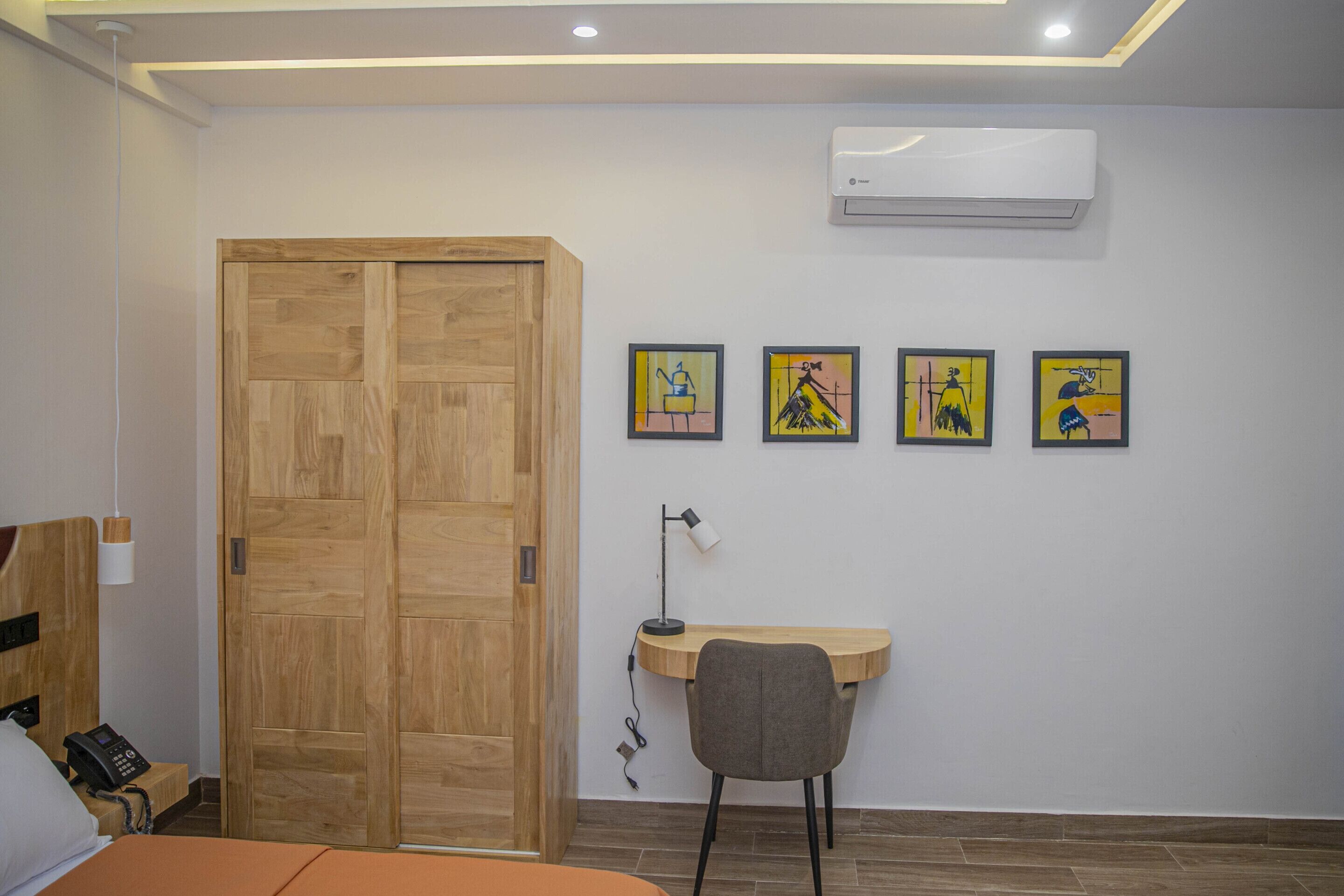 A bedroom with a wooden wardrobe, a desk with a chair, and four framed artworks on the wall.