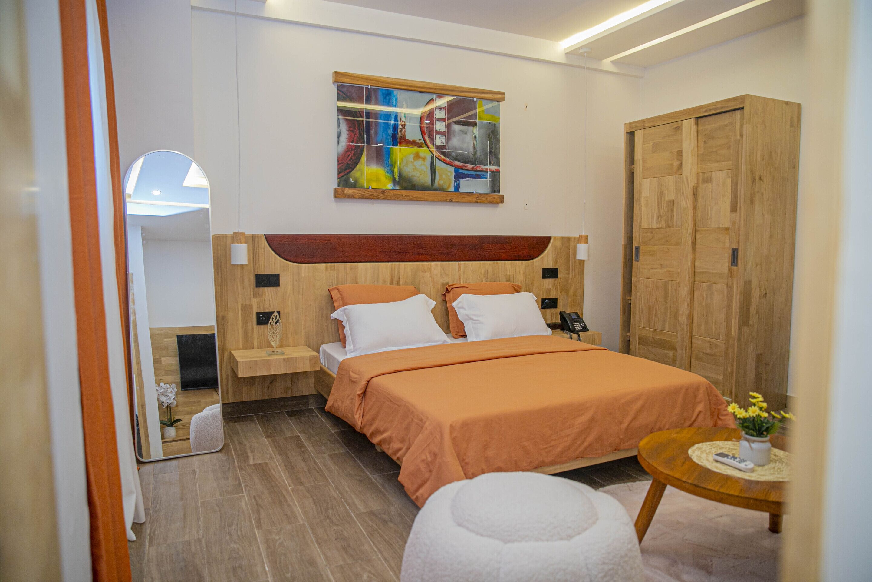 A modern bedroom with a wooden headboard, a large bed with orange bedding, a bedside table with a phone, and a small round table with a vase of flowers.