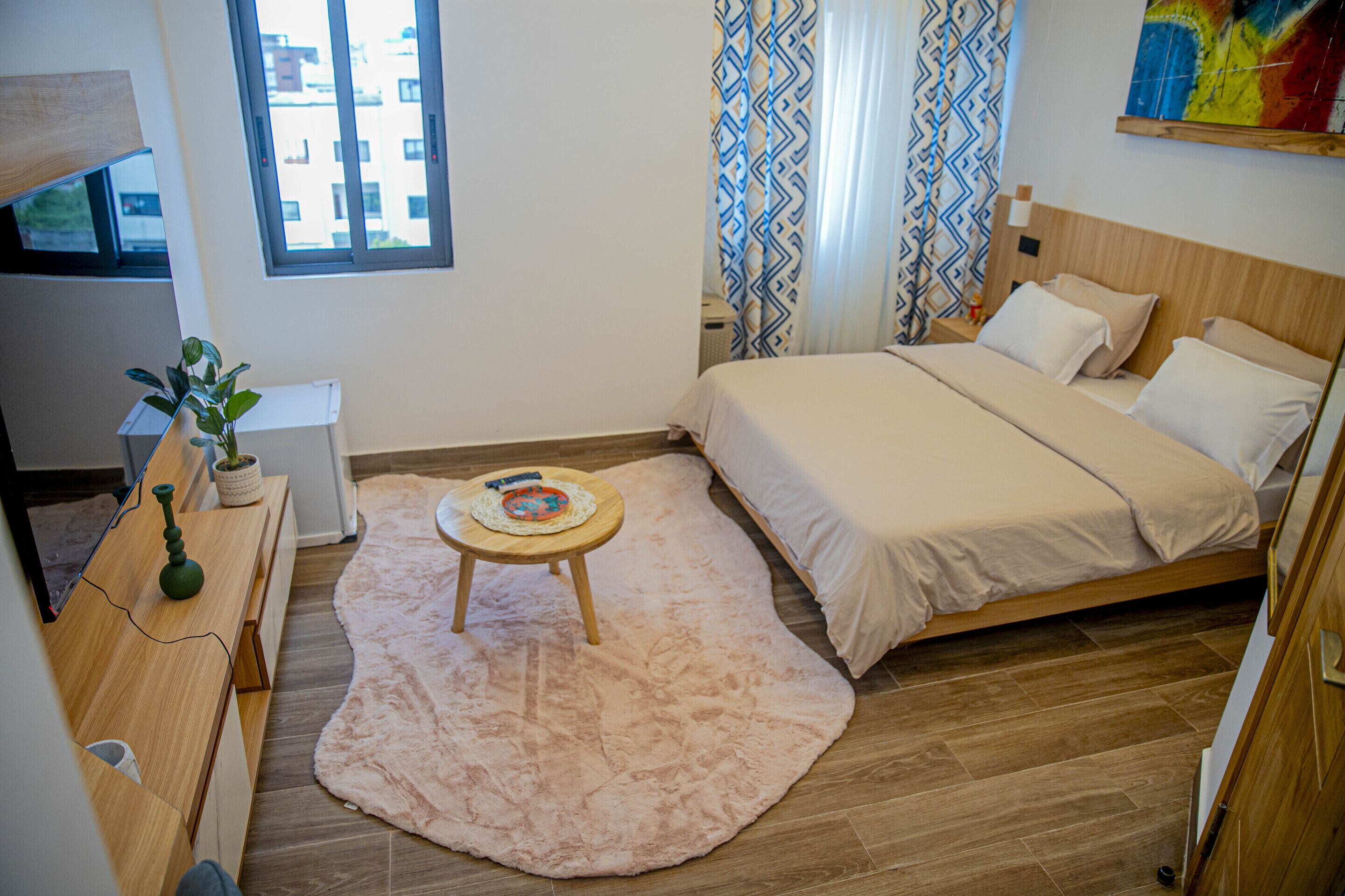 Een slaapkamer met een bed, een houten bureau, een klein tafeltje met een plant, een vloerkleed en een raam met gordijnen.