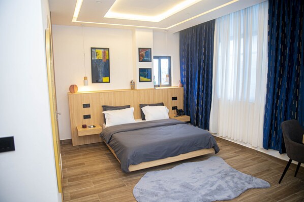 A modern bedroom with a wooden bed, a bedside table, a nightstand, and a large window with blue curtains.