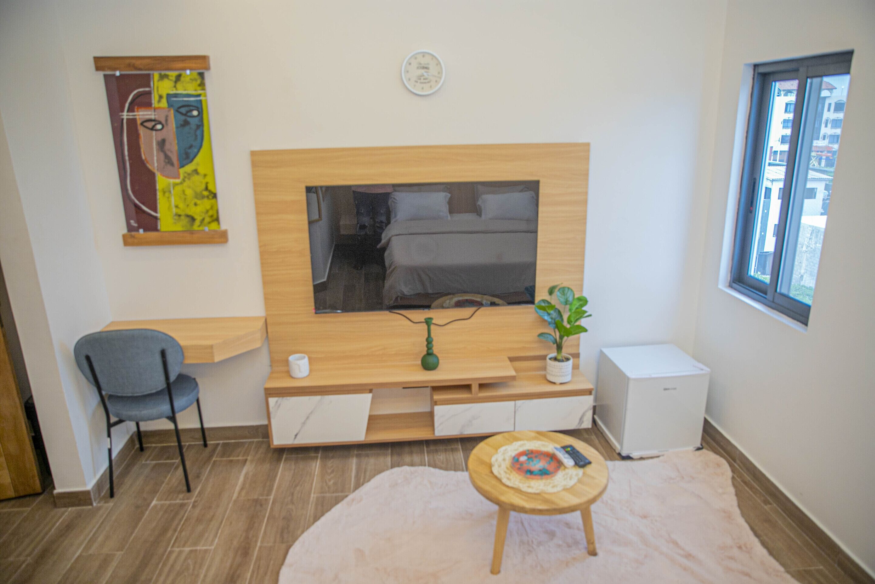 A modern room with a bed, a TV, a desk, a chair, a small fridge, and a window.