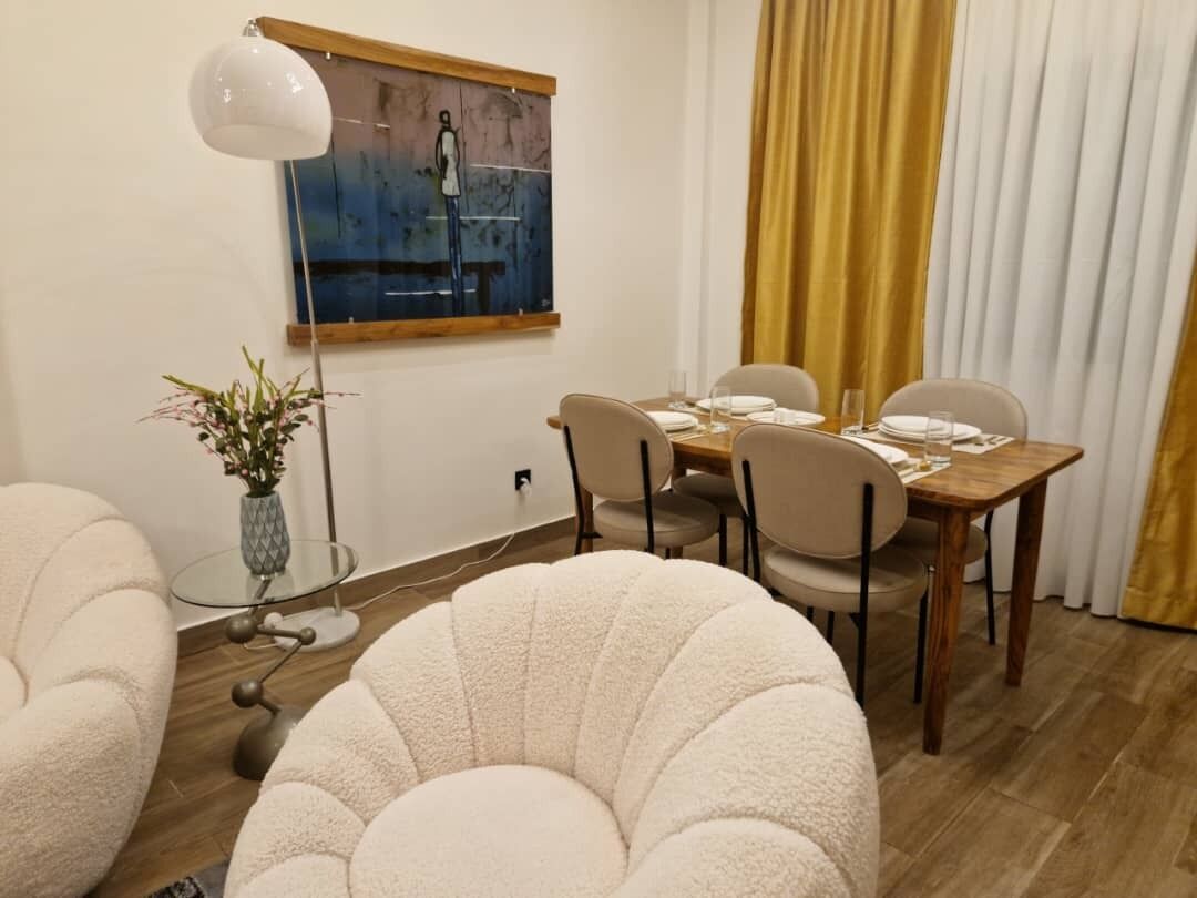 A dining area with a wooden table set for four, surrounded by chairs. There is a glass table lamp on the left, a vase with flowers on a glass table, and a painting on the wall above the table.