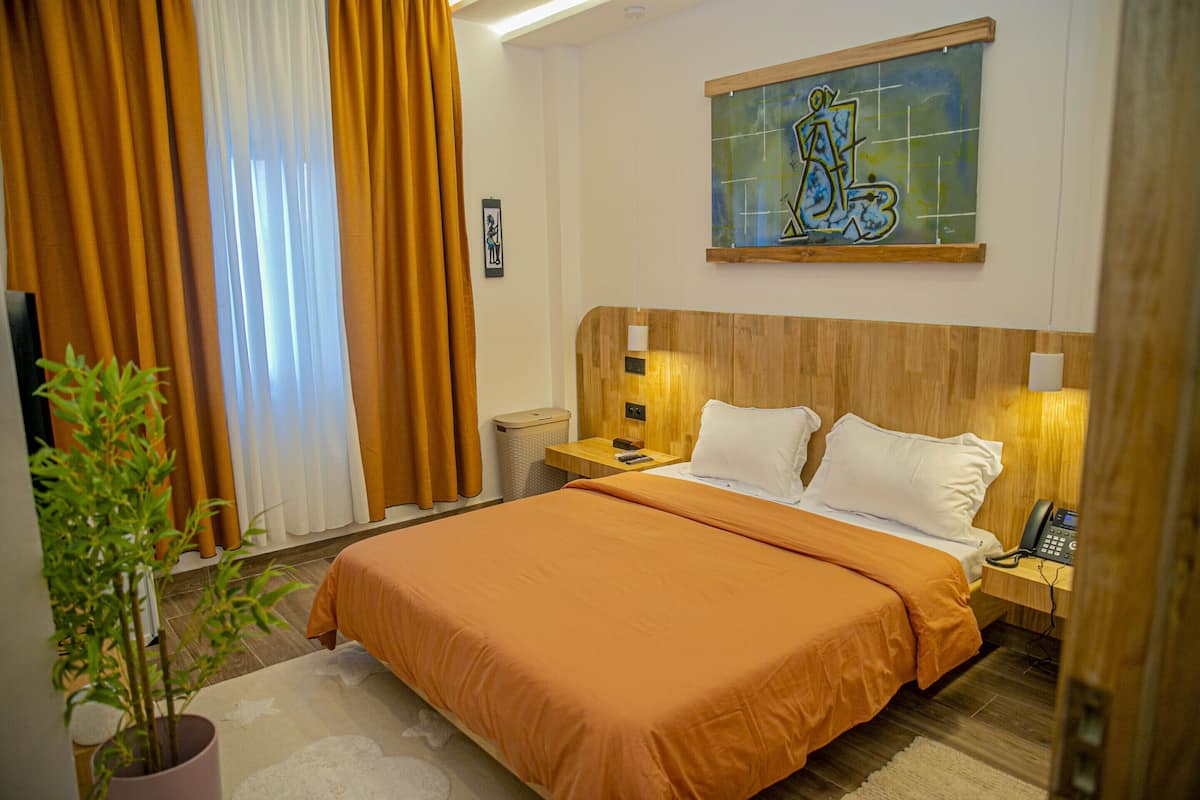 Camera d'albergo con un letto grande, una testiera in legno, comodini con lampade, una pianta in vaso e un quadro appeso al muro.