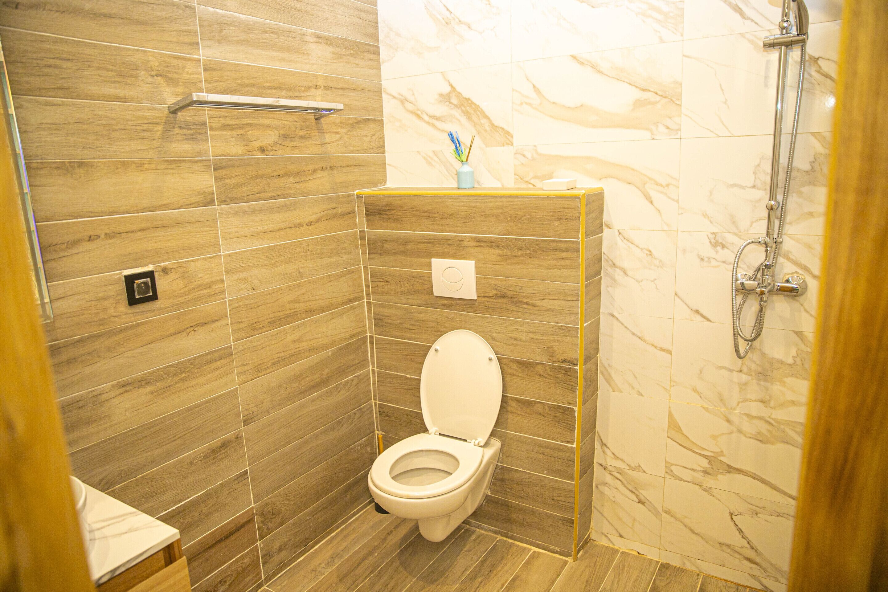 Un baño moderno con piso de madera, inodoro blanco, pared de ducha de mármol y un pequeño jarrón con flores en el mostrador.