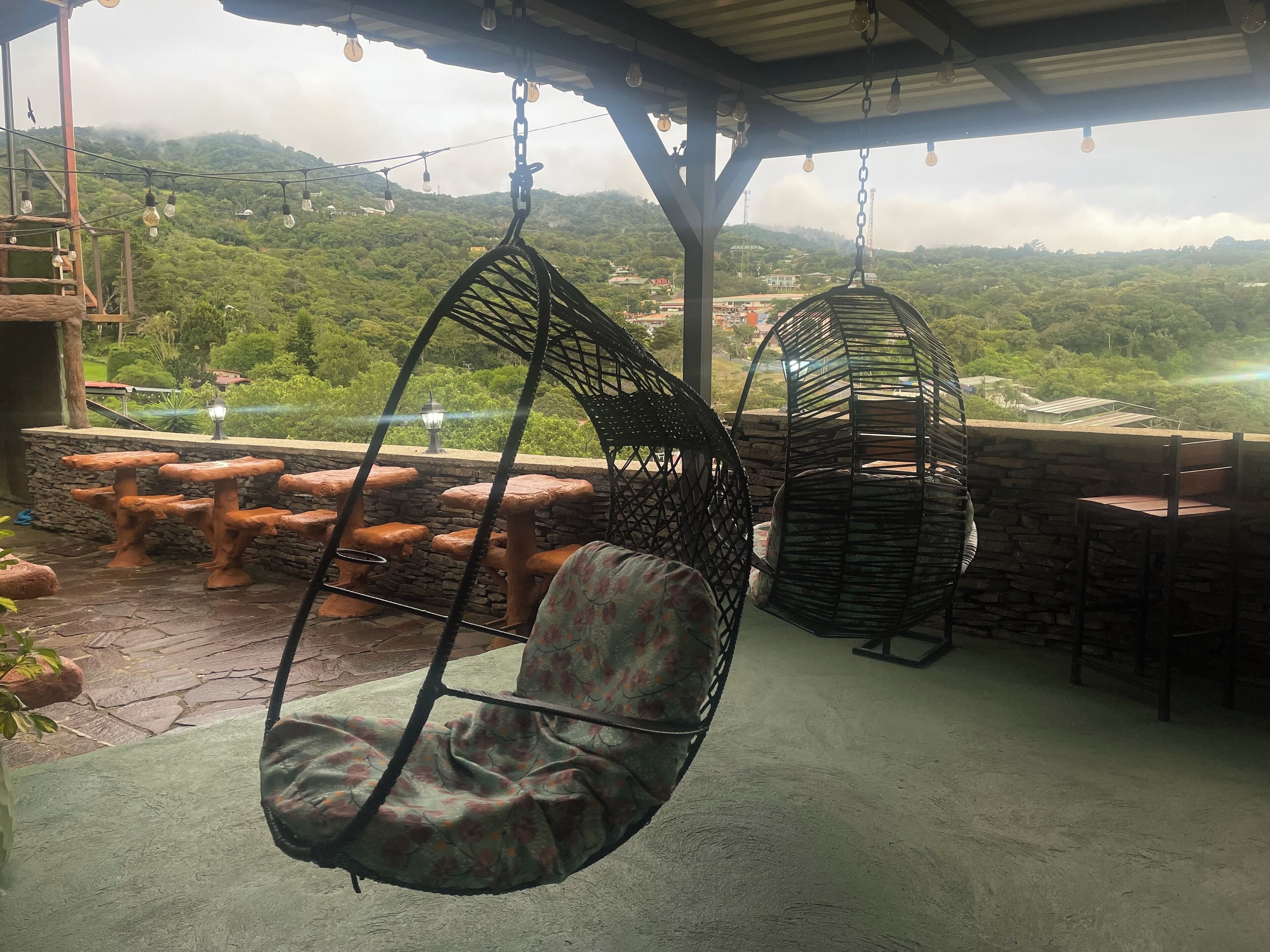 A covered outdoor seating area with hanging chairs and stone tables, overlooking a lush green landscape.
