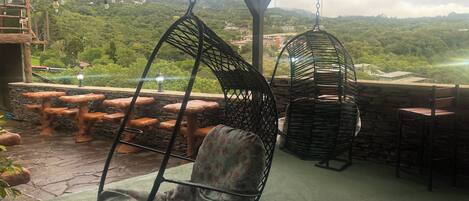 A covered outdoor seating area with hanging chairs and stone tables, overlooking a lush green landscape.