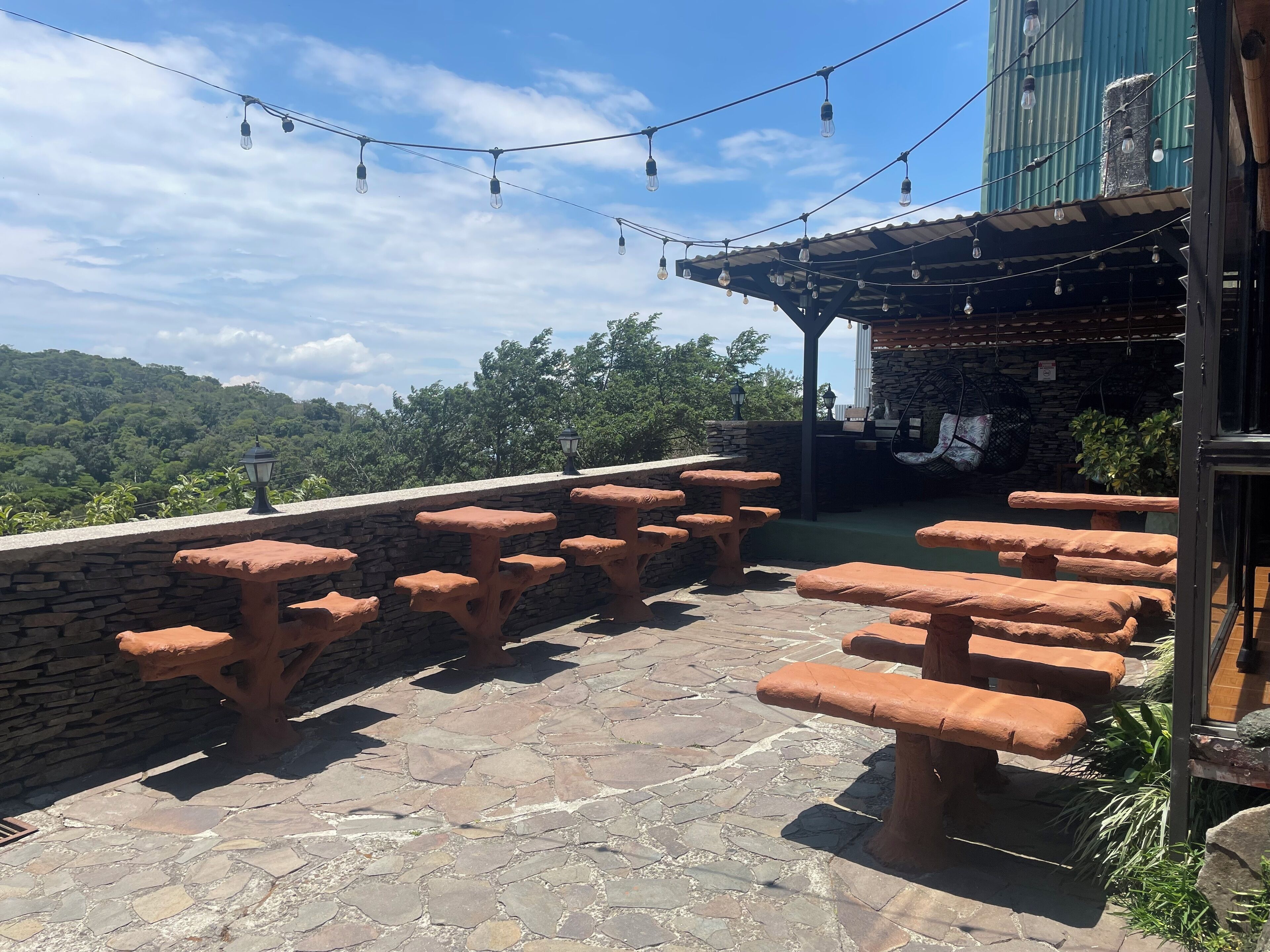 An outdoor patio with stone tables and benches, string lights, and a stone wall.