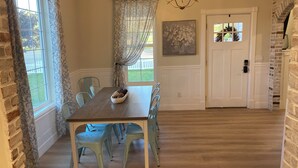 Un coin repas avec une table en bois, des chaises bleu clair, un lustre et une vue sur l’extérieur grâce à une fenêtre ornée de rideaux à motifs.