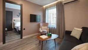 Une chambre d’hôtel moderne avec un canapé, un fauteuil, une table basse avec un livre et un objet vert, un téléviseur fixé au mur et une fenêtre avec des rideaux.