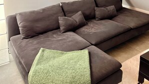 A modern living room with a dark grey sectional sofa, a green throw blanket, and a wooden shelf with decorative items and a framed picture.