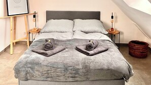 A modern bedroom with a sloped ceiling, a bed with a grey blanket, two bedside tables with lamps, and a framed picture on a wooden easel.