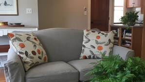 A living room with a grey sofa, patterned cushions, a round table with a fern, a patterned rug, a painting on the wall, and a kitchen area in the background.