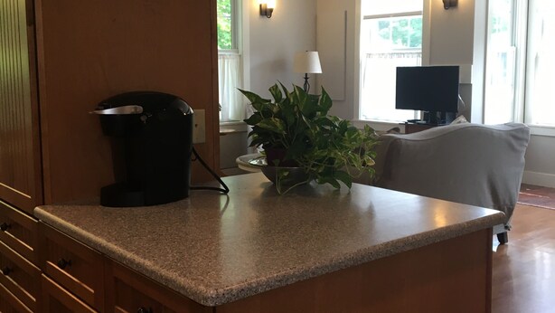 A kitchen with a granite countertop, a coffee maker, and a potted plant. There is a dining area with a chair and a table, and a television in the background.