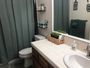 Une salle de bain avec des toilettes, un lavabo, un miroir et une fenêtre avec un rideau. On trouve des plantes dans un panier posé sur le comptoir et une serviette accrochée au mur.