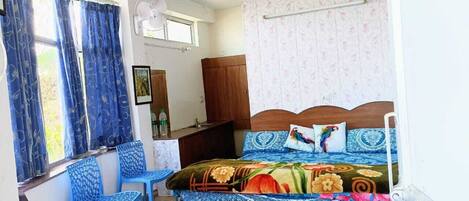 Una camera da letto con un letto, due poltrone blu, un armadio in legno, un ventilatore e una finestra con tende.
