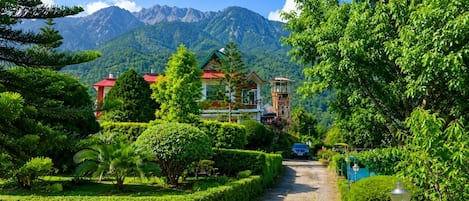A pathway leading to a building with a red roof, surrounded by lush greenery and a mountain backdrop.