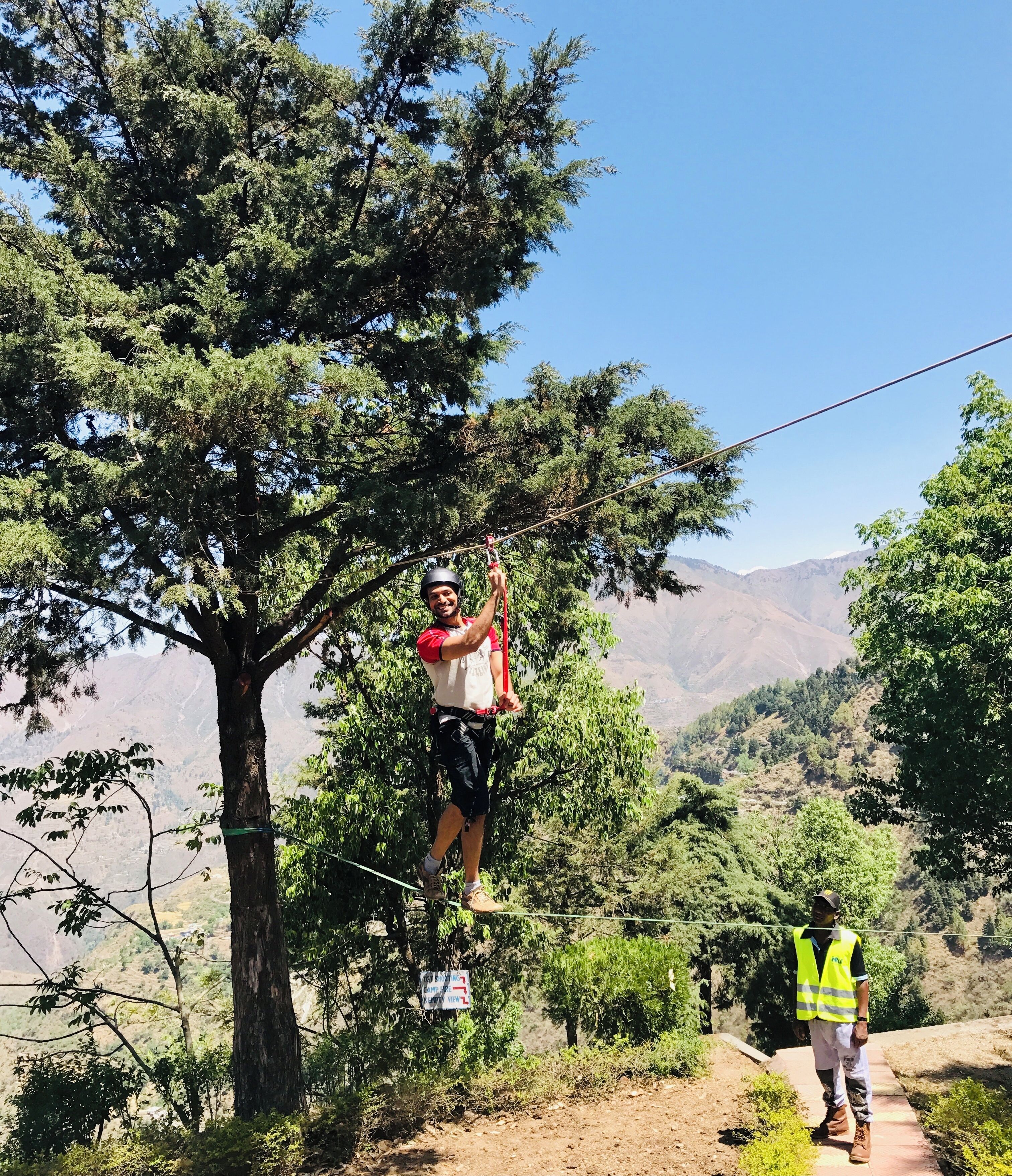 A person on a rope bridge in a natural setting with trees and mountains in the background.