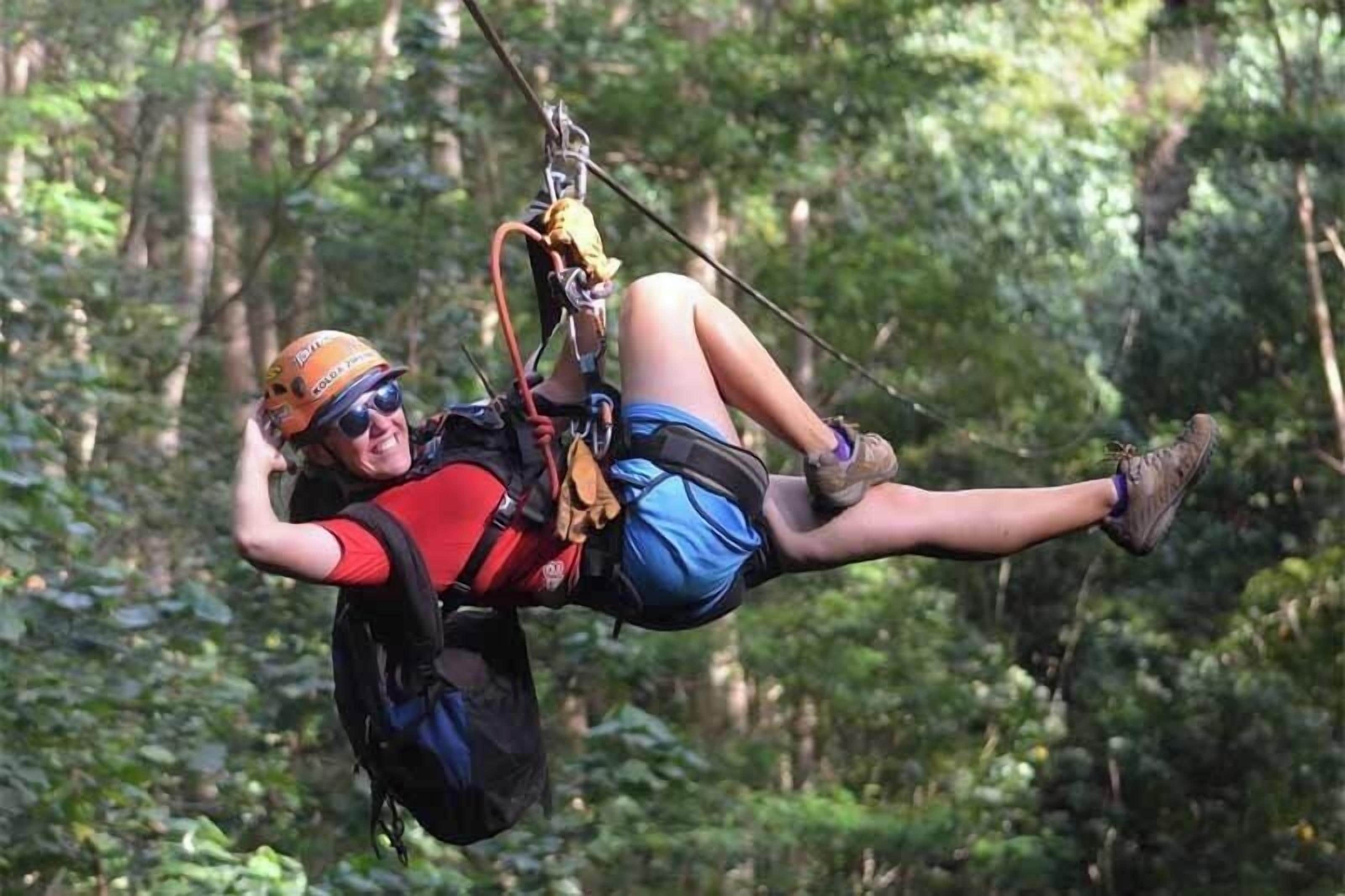 A person suspended in mid-air, wearing a helmet and harness, with a forested background.