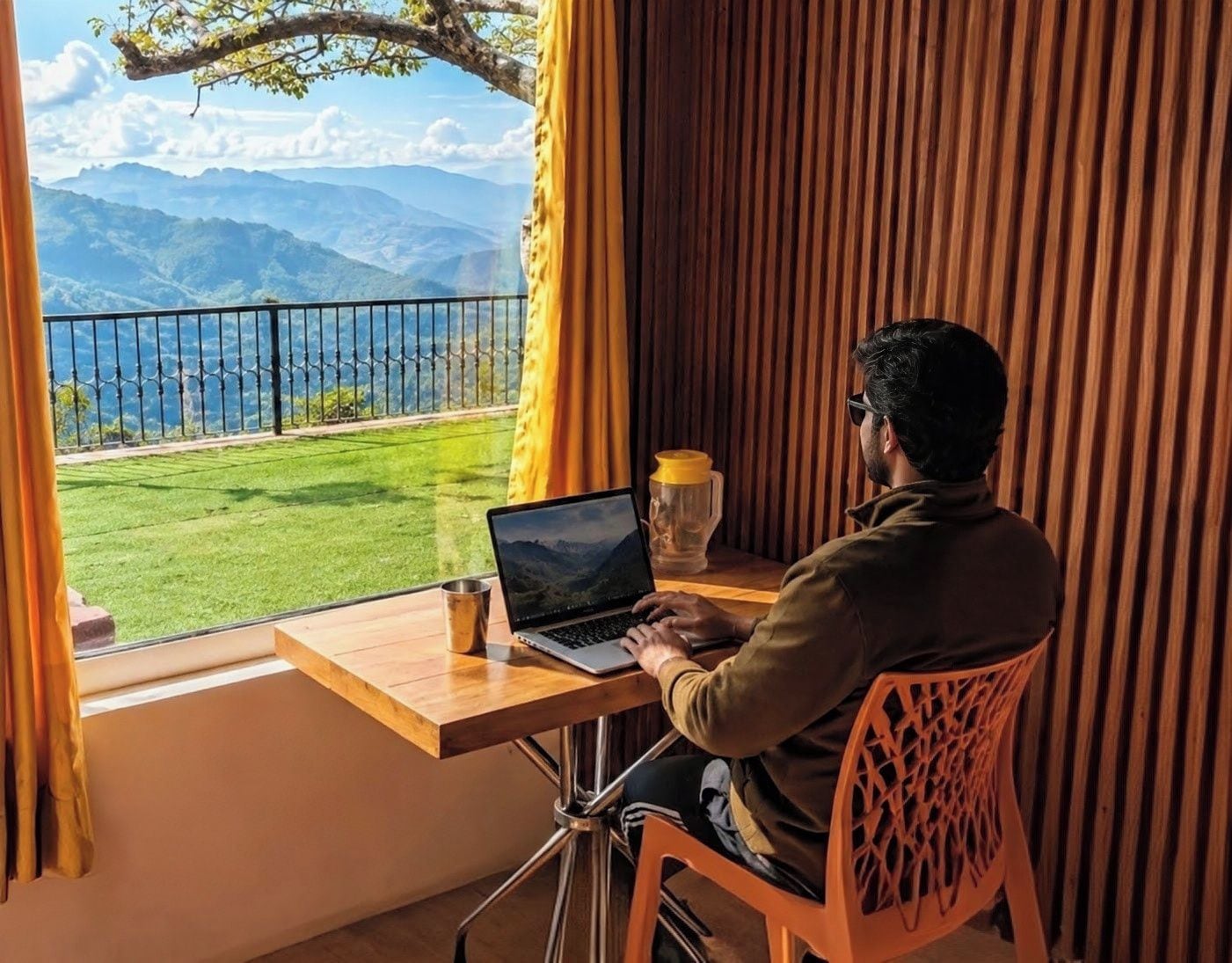 Una persona sentada en una mesa de madera con una computadora portátil, mirando por una ventana con vistas a montañas.