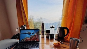 Un portatile su una scrivania di legno con vista panoramica attraverso una finestra, una macchina per caffè e diverse bottiglie.