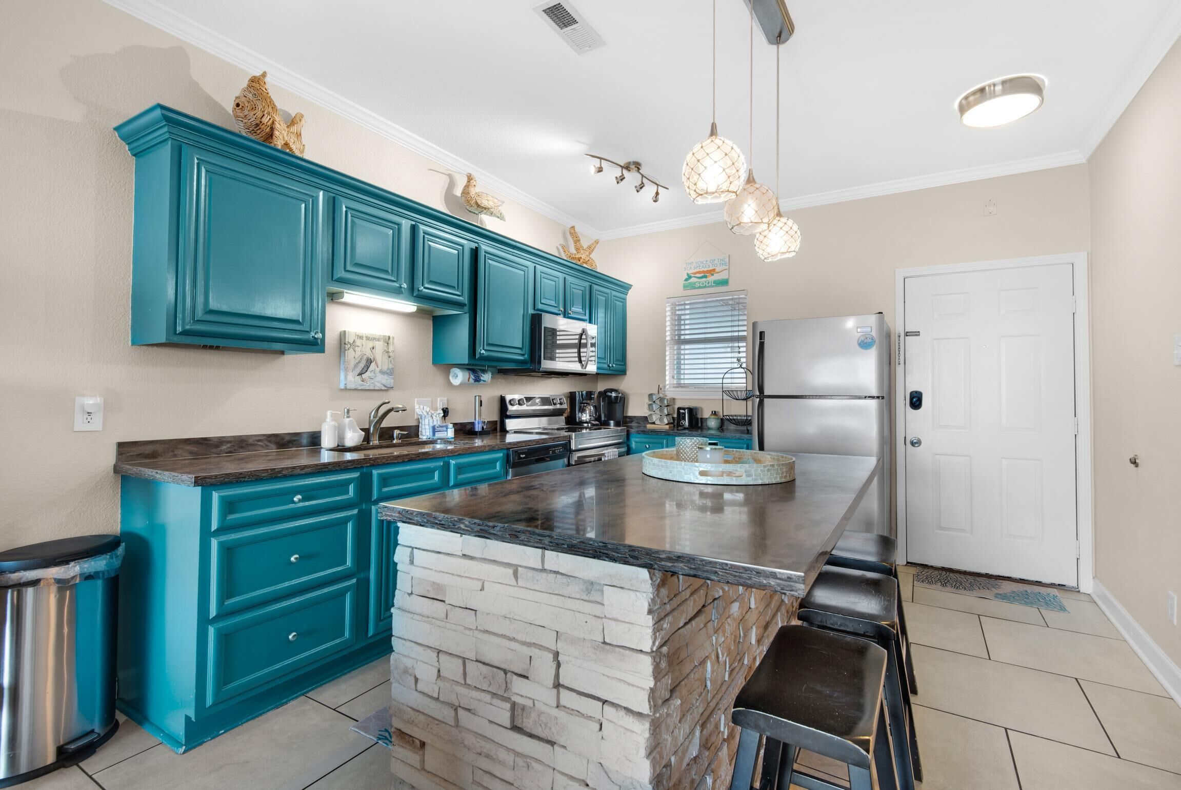 A modern kitchen with teal cabinets, a central island, stainless steel appliances, and pendant lighting.