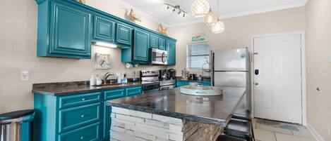 A modern kitchen with teal cabinets, a central island, stainless steel appliances, and pendant lighting.