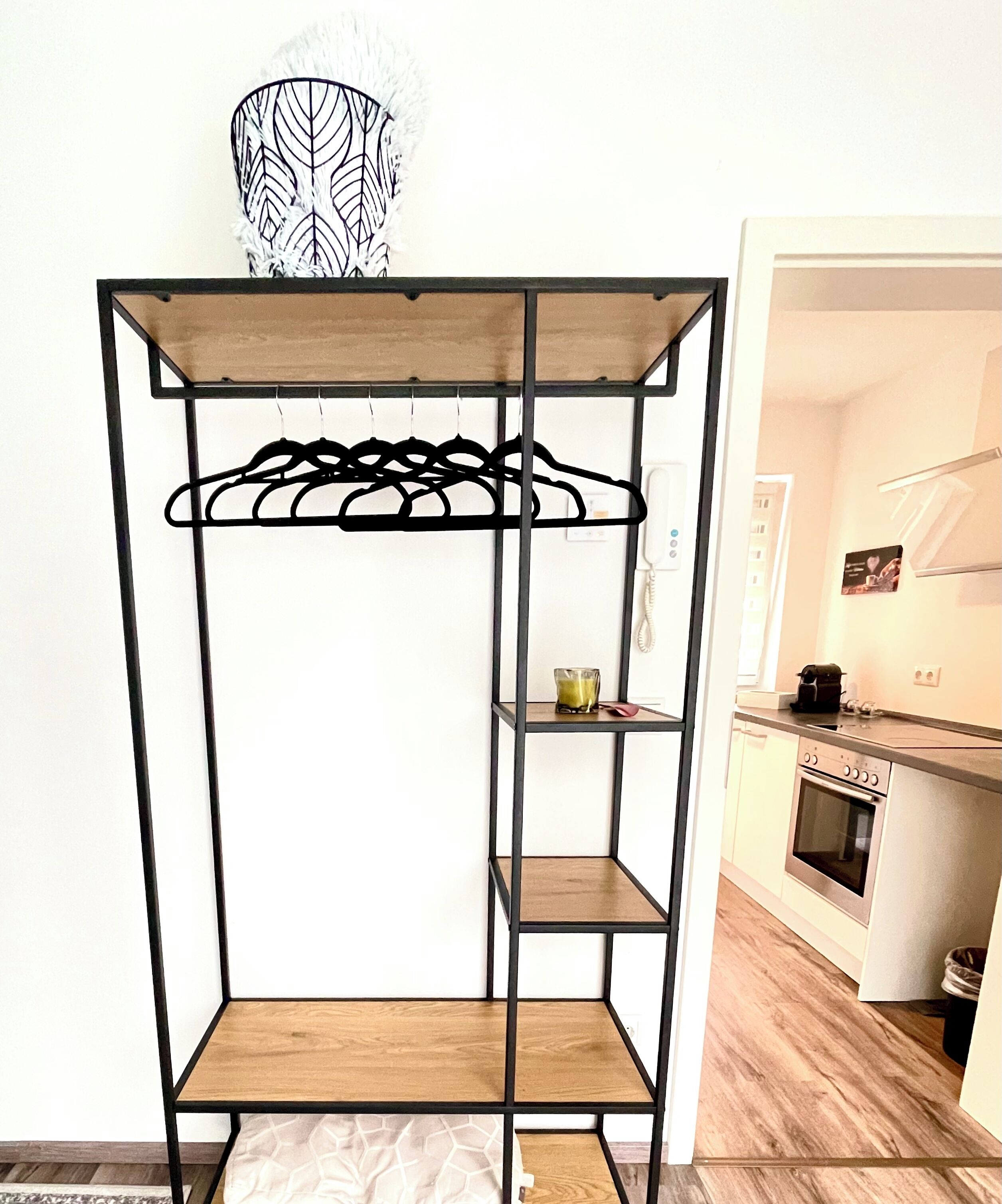 A metal rack with shelves and a hanging section, positioned in a room with a kitchen area visible in the background.