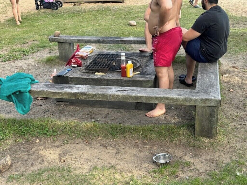 A picnic table with a grill, food items, and a metal bowl on the ground.