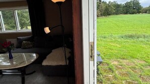 Habitación con sofá, una mesita de vidrio con un florero, una lámpara de pie y vista a un campo verde a través de una puerta abierta.