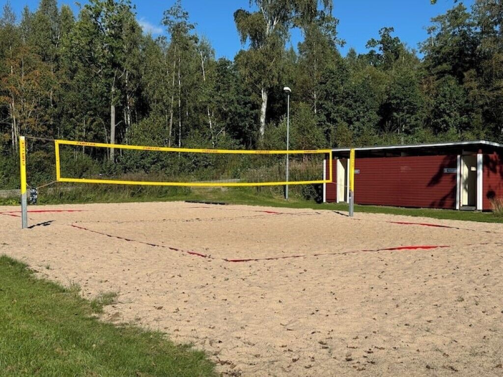 A volleyball court with a net, surrounded by sand and grass, with a red building and trees in the background.
