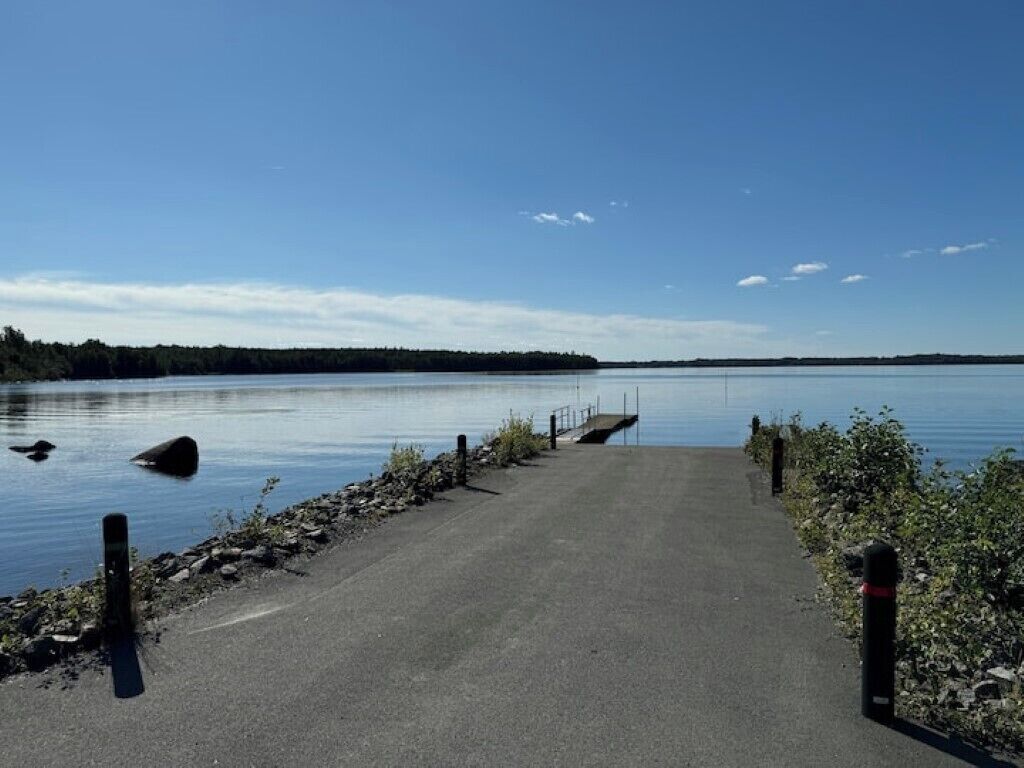 Ein gepflasterter Weg führt zu einem Steg an einem ruhigen See unter blauem Himmel.