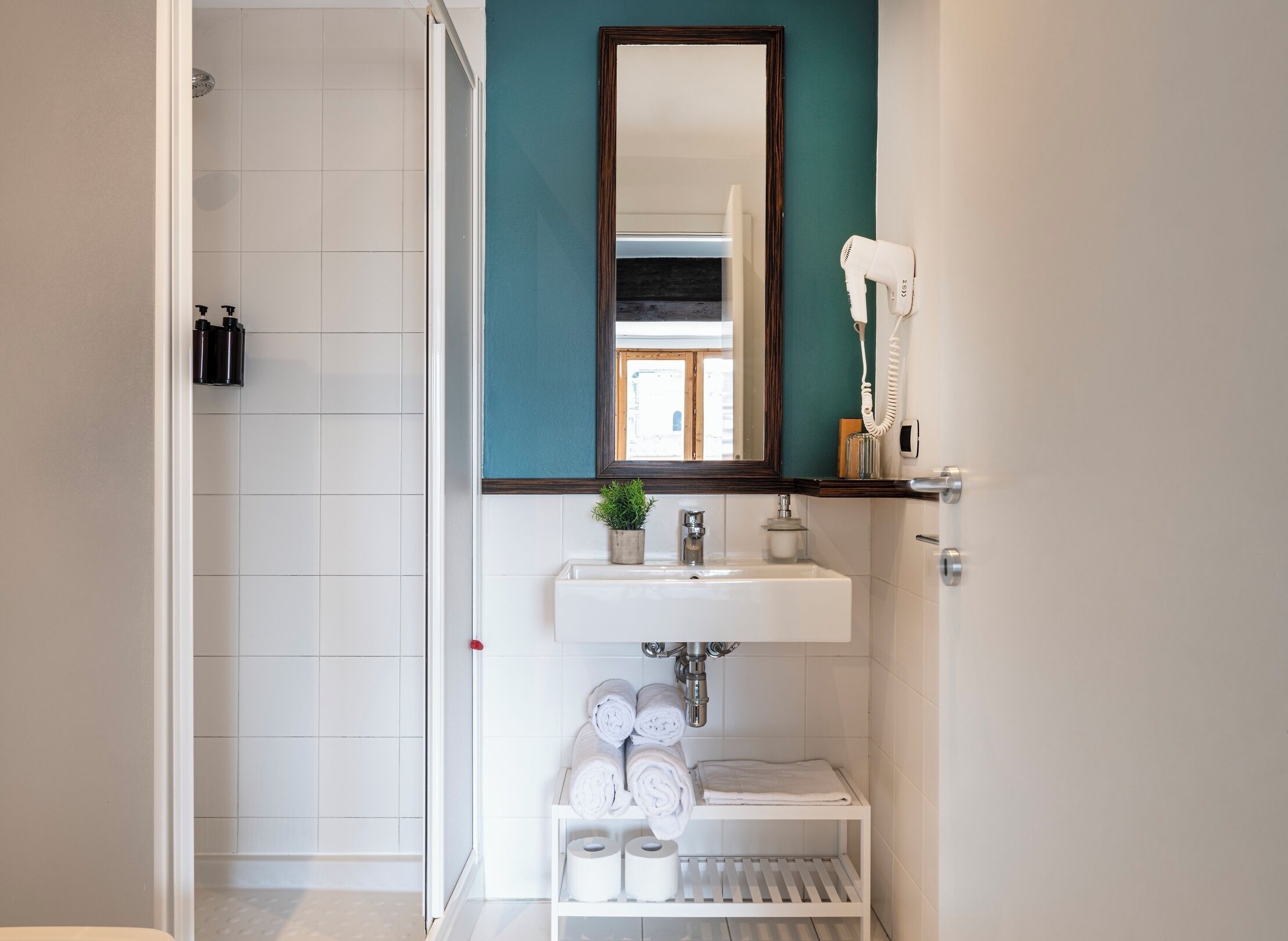 Un baño moderno con un lavamanos blanco, un espejo, un secador de pelo y un estante con toallas.