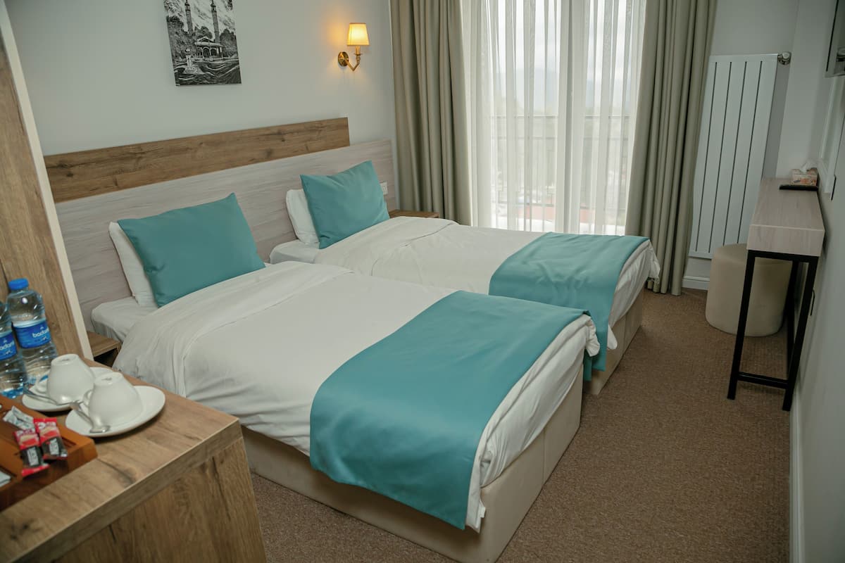 A hotel room with two beds, a wooden headboard, a bedside table with a tray of toiletries, a wall-mounted lamp, and a view of the outdoors through sheer curtains.