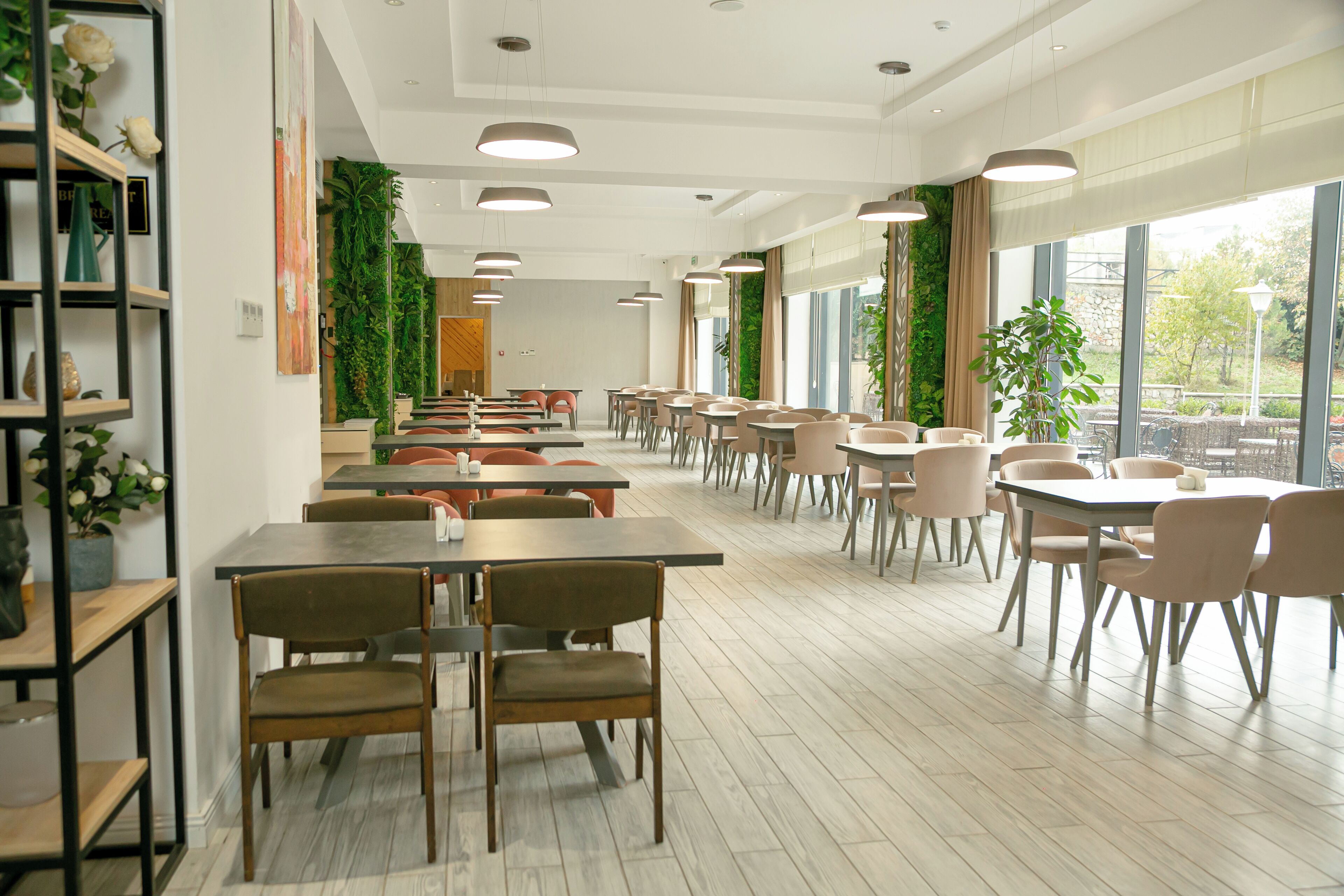 A spacious indoor dining area with multiple tables and chairs, large windows with a view of greenery outside, and a modern ceiling design.