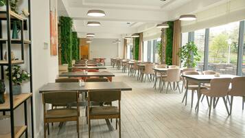 A spacious indoor dining area with multiple tables and chairs, large windows with a view of greenery outside, and a modern ceiling design.