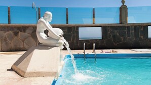 Una estatua junto a una piscina, con un muro de piedra y un cielo azul despejado al fondo.