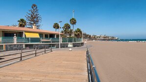Un muelle de madera que conduce a una playa, con un edificio de fachada de piedra y toldo amarillo.