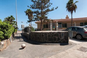A paved pathway leading to a stone wall with a spherical object on it, a parked car, and a building with a tiled roof and green shutters. - Aquarius 18 by VillaGranCanaria (San Bartolomé de Tirajana)