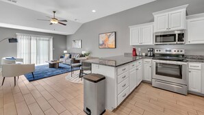 A modern kitchen with white cabinets, a stainless steel oven, and a microwave. There is a dining area with a table and chairs, and a trash can is visible.