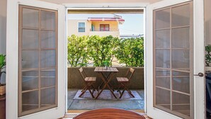 Una puerta abierta que da a un balcón con una mesa y sillas, una planta en maceta, y un edificio al fondo con toldo rojo y blanco.