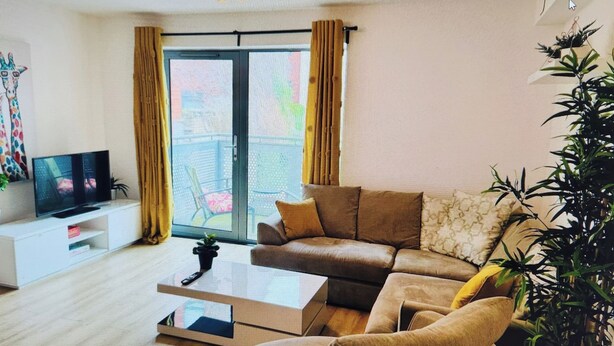 Una sala moderna con un sofá, una mesa de centro y un televisor sobre un soporte blanco. Hay un ventanal corredizo que da a un área exterior con sillas.