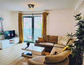 Una sala moderna con un sofá, una mesa de centro y un televisor sobre un soporte blanco. Hay un ventanal corredizo que da a un área exterior con sillas.