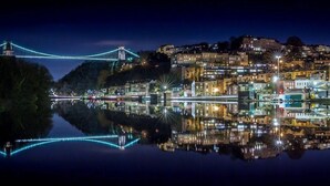 Un paisaje urbano nocturno con un puente iluminado que se refleja en el agua, edificios con luces y una colina al fondo.
