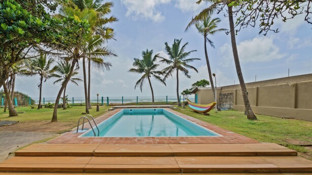 A rectangular swimming pool with a wooden deck in the foreground, surrounded by palm trees and a view of the ocean. - Amy's Haven (Wadduwa)