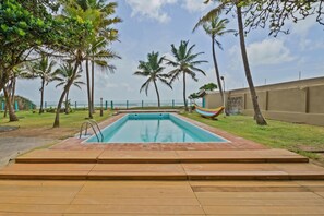 A rectangular swimming pool with a wooden deck in the foreground, surrounded by palm trees and a view of the ocean.